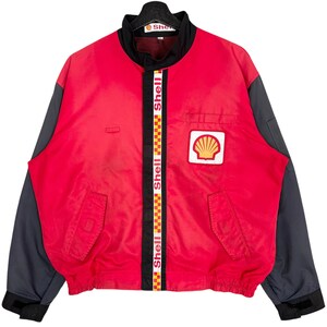 Puede incluir: Chaqueta de carreras Shell roja y gris. La chaqueta tiene un cuerpo rojo, mangas grises y un cuello negro. La parte delantera presenta un parche con el logo de Shell y una franja vertical con el logo de Shell. Tiene dos bolsillos delanteros.