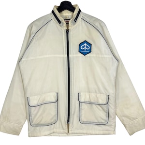 Puede incluir: Chaqueta color crema con cremallera completa, dos bolsillos delanteros y cuello alzado. La chaqueta presenta ribetes azul marino y un parche con el logotipo de Piaggio azul y blanco en el pecho. Chaqueta ligera de estilo vintage.