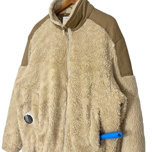 May include: A beige sherpa jacket with brown fabric panels on the shoulders and arms. The jacket has a zipper closure and a pocket on the left side. A blue tool is sticking out of the pocket.