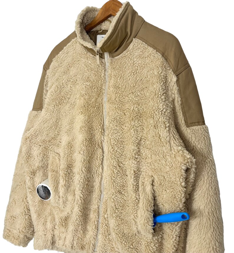 May include: A beige sherpa jacket with brown fabric panels on the shoulders and arms. The jacket has a zipper closure and a pocket on the left side. A blue tool is sticking out of the pocket.
