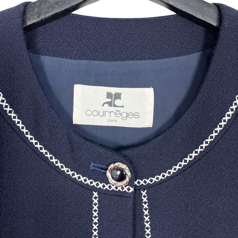 May include: A navy blue blazer with a white stitched trim around the collar and button placket. The blazer has a single button closure and features a white label with the text "Courr&egrave;ges Paris" on the front.