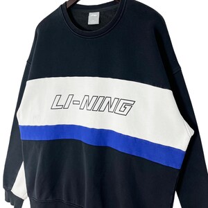 May include: Black sweatshirt with a white and blue stripe design. The word "LI-NING" is printed in white on the white stripe.