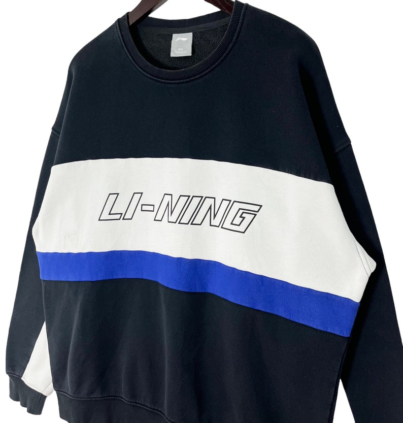 May include: Black sweatshirt with a white and blue stripe design. The word "LI-NING" is printed in white on the white stripe.