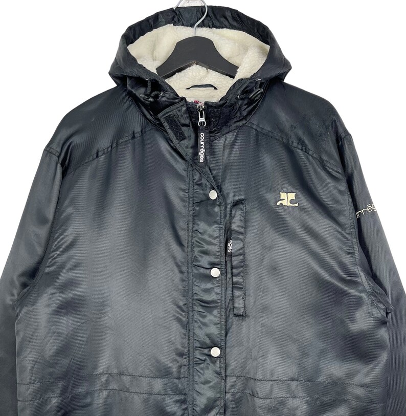 May include: Black hooded jacket with a fleece lining. The jacket has a zipper closure and a button placket. The jacket has a logo on the left chest and the word "Courage" on the zipper.