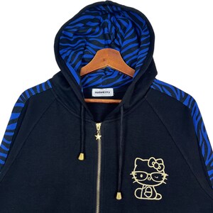 May include: Black zip-up hoodie with blue and black zebra print on the sleeves and hood lining. The hoodie features a gold Hello Kitty graphic with glasses.