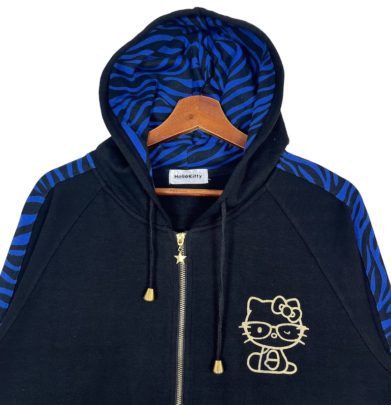 May include: Black zip-up hoodie with blue and black zebra print on the sleeves and hood lining. The hoodie features a gold Hello Kitty graphic with glasses.