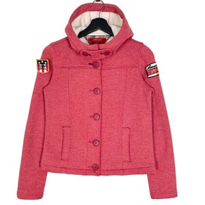 May include: A pink hooded jacket with a button closure and two patch pockets. The jacket has two embroidered patches, one with the text "Tommy Girl" and the other with the text "American Tommy Girl".