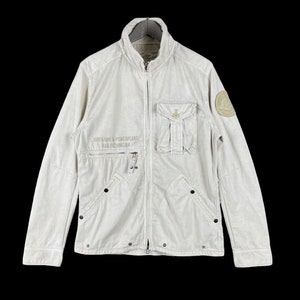 Può includere: Giacca bianco sporco con zip, due tasche sul petto e una piccola tasca sulla manica sinistra. La giacca ha il testo "AIRFRAME & POWERPLANT FAA TECHNICIAN" stampato sul davanti. Una toppa circolare è sulla manica destra.