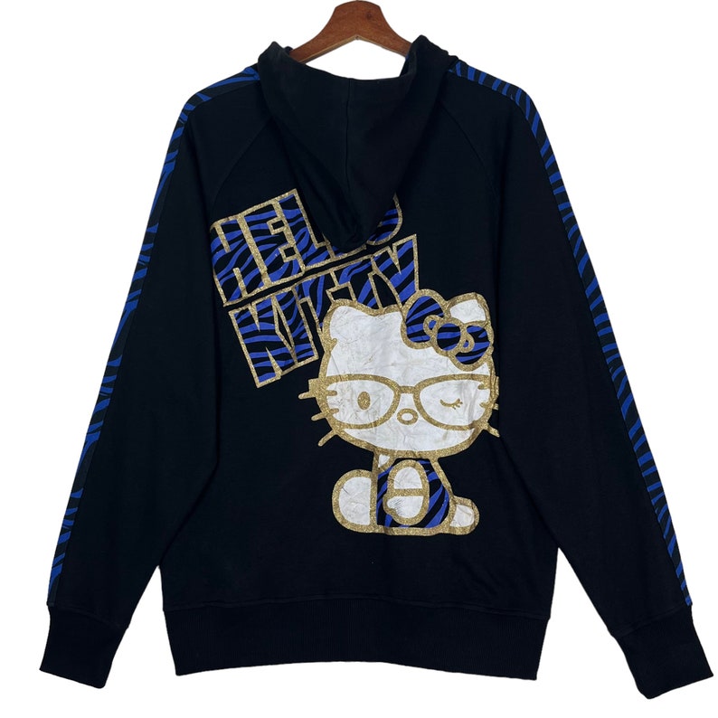 May include: Black hooded sweatshirt with a blue and gold Hello Kitty graphic on the back. The graphic features Hello Kitty wearing glasses and a headband with a bow.