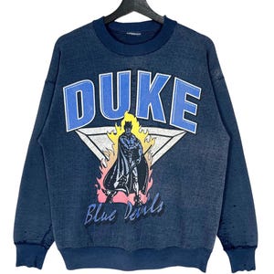 Może przedstawiać: Granatowa bluza z nadrukiem vintage Duke Blue Devils. Grafika przedstawia kreskówkowego diabła w pelerynie z płomieniami za nim. Tekst "DUKE" znajduje się nad grafiką, a "Blue Devils" pod nią.