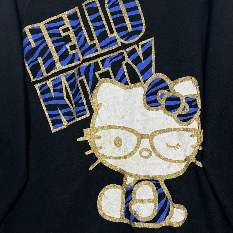 May include: Black sweatshirt with a gold and blue "Hello Kitty" graphic. The graphic features Hello Kitty wearing glasses and a blue and gold zebra print bow.