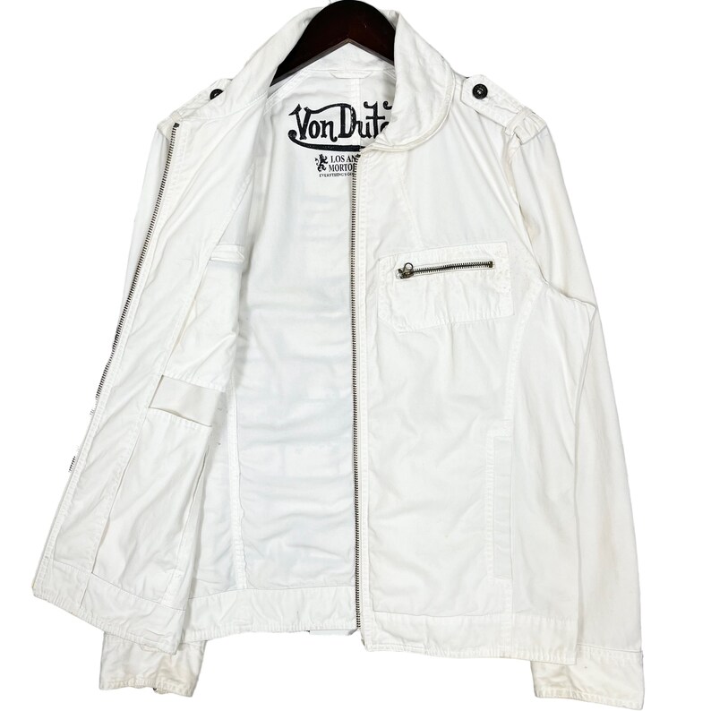 K&ouml;nnte beinhalten: Wei&szlig;e Rei&szlig;verschlussjacke mit einem Von Dutch Logo auf der Vorderseite. Die Jacke hat einen Stehkragen und zwei Taschen auf der Vorderseite.