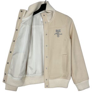 May include: Cream-colored varsity jacket with a white ribbed collar, cuffs, and hem. The jacket features a button-up front, a pocket, and a silver emblem on the chest. The interior lining is white.
