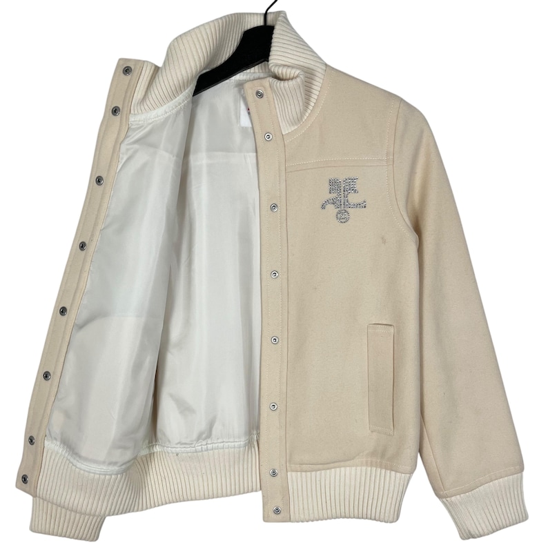 May include: Cream-colored varsity jacket with a white ribbed collar, cuffs, and hem. The jacket features a button-up front, a pocket, and a silver emblem on the chest. The interior lining is white.