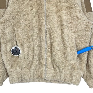 May include: A beige fleece jacket with a zipper closure and two pockets. The jacket has a brown trim on the shoulders and sleeves.
