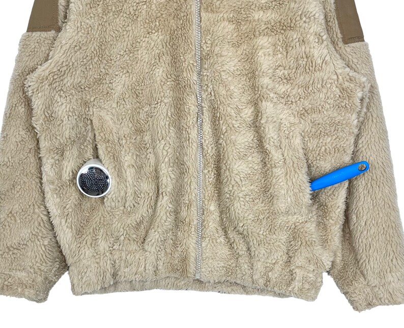 May include: A beige fleece jacket with a zipper closure and two pockets. The jacket has a brown trim on the shoulders and sleeves.