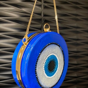 Pu&ograve; includere: Una pochette rotonda blu con una tracolla a catena dorata. La pochette presenta un grande disegno a occhio con dettagli bianchi, blu e neri. La chiusura e la catena sono dorate.