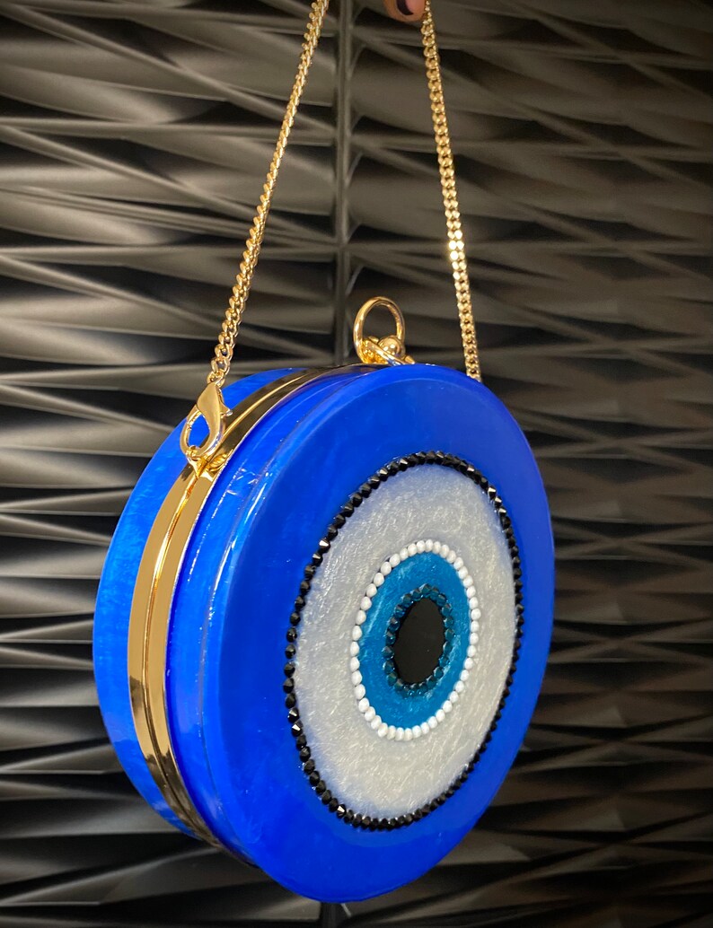 Pu&ograve; includere: Una pochette rotonda blu con una tracolla a catena dorata. La pochette presenta un grande disegno a occhio con dettagli bianchi, blu e neri. La chiusura e la catena sono dorate.