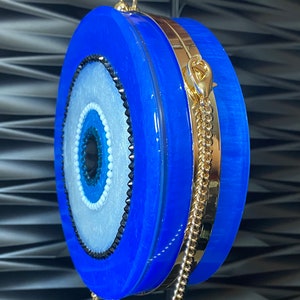 Pu&ograve; includere: Una pochette rotonda blu con una tracolla e dettagli dorati. La pochette presenta un design a occhio centrale con dettagli bianchi, azzurri e neri. La borsa &egrave; tenuta da una mano.