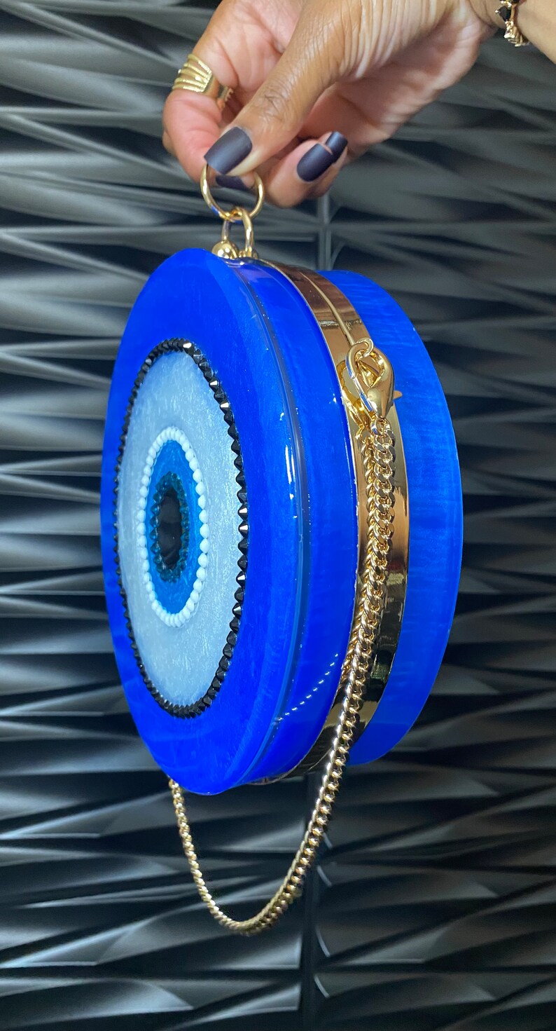 Pu&ograve; includere: Una pochette rotonda blu con una tracolla e dettagli dorati. La pochette presenta un design a occhio centrale con dettagli bianchi, azzurri e neri. La borsa &egrave; tenuta da una mano.