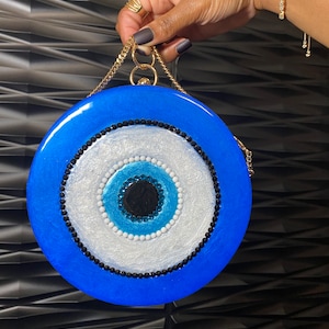 Pu&ograve; includere: Una pochette rotonda blu con una tracolla a catena dorata. La pochette presenta un grande design a occhio stilizzato con una pupilla nera, circondata da accenti bianchi, azzurri e neri. La borsa &egrave; tenuta da una mano con gioielli d'oro.