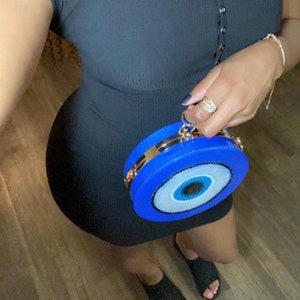 Pu&ograve; includere: Un miniabito nero a coste con scollo a V e maniche corte. Una borsa a mano circolare blu brillante con una tracolla a catena dorata e un grande disegno a occhio &egrave; tenuta in mano. La borsa ha un bordo dorato.
