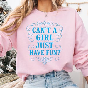Puede incluir: Sudadera rosa claro con el texto azul "CAN'T A GIRL JUST HAVE FUN?". La sudadera se combina con vaqueros de mezclilla azul claro. La imagen está ambientada en un ambiente festivo y navideño.