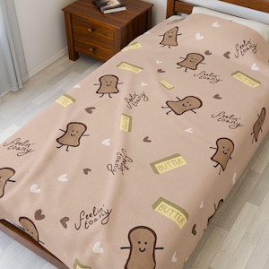 May include: A beige bedspread featuring a repeating pattern of cartoon toast slices, butter blocks, and hearts. The words "Feelin' Toasty" are written in a cursive font. The bedspread is on a wooden bed frame.