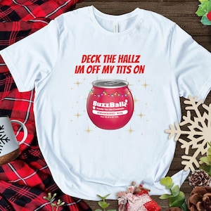 Może przedstawiać: Biała koszulka z czerwonym napisem "Deck the Hallz Im Off My Tits On". Koszulka przedstawia grafikę czerwonej puszki BuzzBall z łańcuchem świetlnym. W tle kubek z motywem płatka śniegu i czerwono-czarny wzór w kratę.