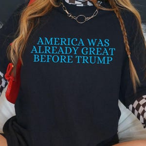 Może przedstawiać: Czarny t-shirt z napisem "AMERICA WAS ALREADY GREAT BEFORE TRUMP" w kolorze niebieskim. Osoba nosi srebrny naszyjnik z łańcuszkiem i koszulę z długim rękawem w szachownicę pod spodem. Widoczna jest czerwona torebka.