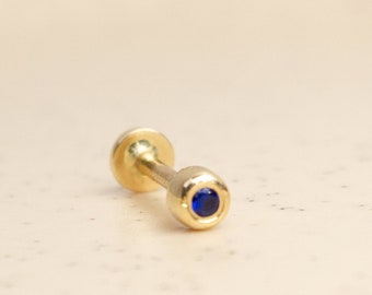 Puede incluir: Un pendiente de botón de color dorado con una pequeña gema redonda de color azul oscuro en un extremo. El pendiente tiene una parte posterior plana y un poste para perforar. El diseño es sencillo y elegante, adecuado para el uso diario. Mide aproximadamente 1 cm de largo.