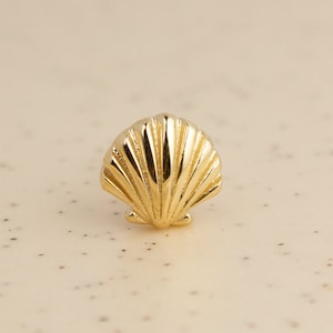 May include: A small, gold-colored seashell. The shell has a detailed, ridged surface and is set against a neutral background. The design is likely for jewelry or decorative purposes.