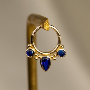 May include: Gold hoop earring with a teardrop-shaped blue gemstone dangling from the bottom and two smaller blue gemstones on either side.