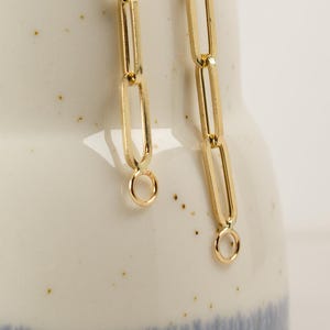 Op de afbeelding: Twee gouden kettingen met een schakelstijl. De kettingen hangen aan een witte keramische vaas met een blauw-wit gespikkeld ontwerp.
