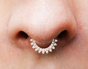 May include: A close-up of a person's nose with a gold septum ring adorned with small, clear rhinestones.