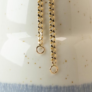 May include: Two gold chains with jump rings hanging from a white ceramic vase with a blue and brown glaze.