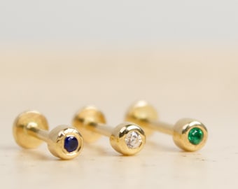 Puede incluir: Tres pendientes de botón dorados, cada uno con una gema de diferente color: un zafiro azul oscuro, un diamante transparente y una esmeralda verde. Se muestran sobre una superficie clara.