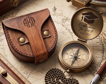 May include: A brass pocket compass with a chain, featuring an open lid engraved with a graduation cap, the text "Congratulaions You Did It", and the date "06.08.2027". Beside it sits a brown leather pouch with the monogram "AKA" and brass snaps.