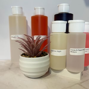 Könnte beinhalten: Eine Sammlung klarer Plastikflaschen mit weißen Deckeln, einige gefüllt mit farbigen Flüssigkeiten. Vorne steht ein kleiner weißer Topf mit einer Sukkulentenpflanze. Auf den Etiketten der Flaschen steht "Egyptian Night" und "Freesia & Peppermint Blossom".