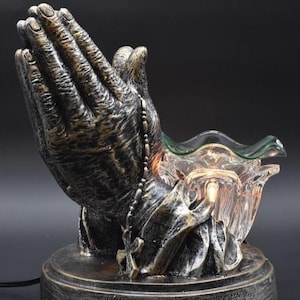 Könnte beinhalten: Ein dekorativer elektrischer Ölverdampfer in Form von betenden Händen. Die Hände sind bronzefarben mit strukturierter Oberfläche und halten eine klare Glasschale. Ein Rosenkranz hängt zwischen den Händen. Der Sockel hat eine Inschrift.