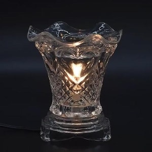 Puede incluir: Un calentador de aceite de vidrio transparente con un patrón de corte de diamante. El calentador tiene un borde festoneado y una base con un pie pequeño y redondo. Una luz cálida brilla desde adentro, iluminando el intrincado diseño.
