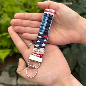May include: A fabric wristlet keychain featuring a patriotic pattern with navy blue, white stars, and red stripes. The strap is attached to a silver-toned metal clasp and a circular split ring. A small beige fabric tag reads Made by Krista Marie.