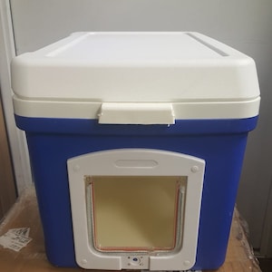 May include: A blue and white cooler with a pet door cut out. The door has a white frame and a clear plastic flap.