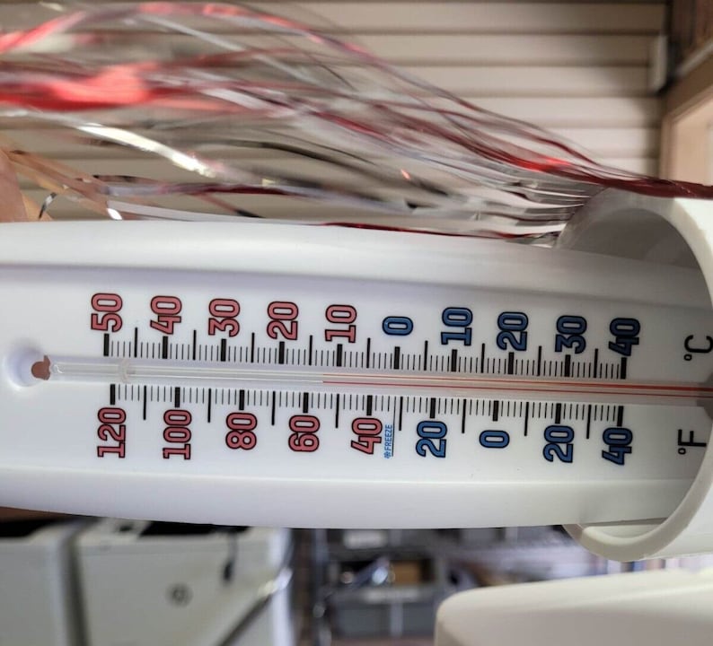 May include: A white thermometer showing both Celsius and Fahrenheit scales. The temperature reads approximately 4&deg;C and 40&deg;F. The thermometer has red and blue markings, with a red liquid column indicating the current temperature. The background is blurred.