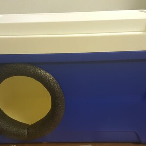 May include: A blue and white cooler with a round opening covered in black foam. The cooler is a repurposed item used as a pet bed.