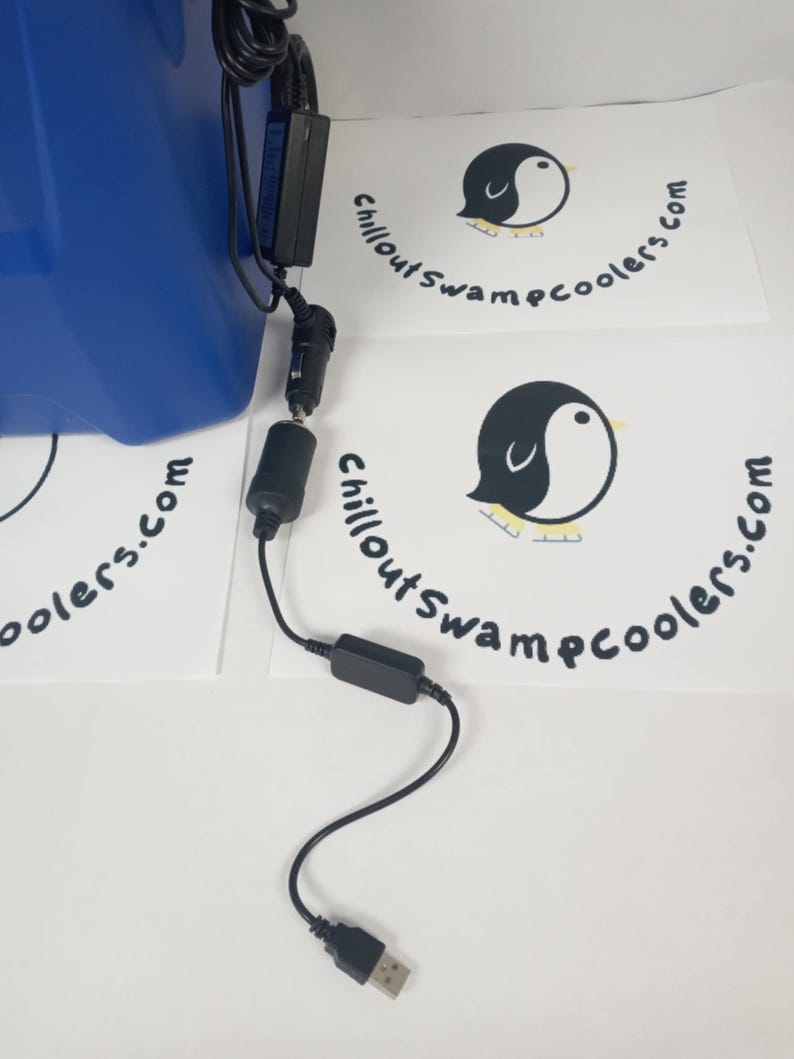 Peut inclure: Un cordon d'alimentation noir avec un connecteur USB, un adaptateur de voiture et un petit bo&icirc;tier noir. Le cordon est connect&eacute; &agrave; une glaci&egrave;re bleue. Le texte "ChilloutSwampcoolers.com" est imprim&eacute; sur du papier blanc.