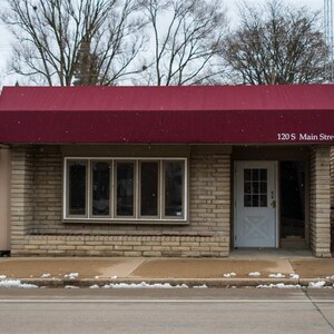 May include: A brick building with a red awning and a white door. The building has a large window with multiple panes. The awning has the text "120 S Main Street" on it.