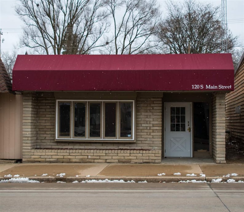 May include: A brick building with a red awning and a white door. The building has a large window with multiple panes. The awning has the text "120 S Main Street" on it.