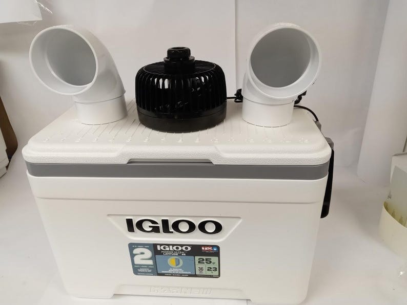 Puede incluir: Una nevera blanca Igloo con un ventilador negro y dos tubos de ventilaci&oacute;n blancos en la parte superior. La nevera tiene una pantalla digital de temperatura que muestra 25 grados Fahrenheit. La nevera est&aacute; etiquetada como "Igloo Ultra 25 Qt."