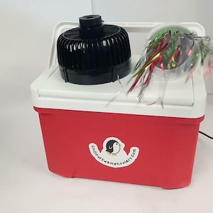 May include: Red and white cooler with a black fan on top. A clear container with colorful streamers sits on the cooler. The cooler has a white handle and a sticker with a penguin logo and the text "chilloutswampcoolers.com".